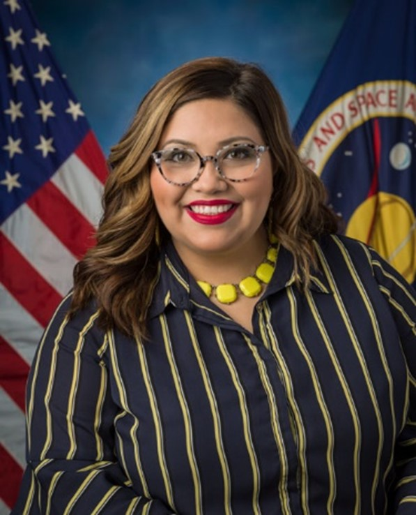 A person wearing a striped brown and yellow blouse smiles in front of a blue background with the United States, left, and NASA, right, flags behind them. 